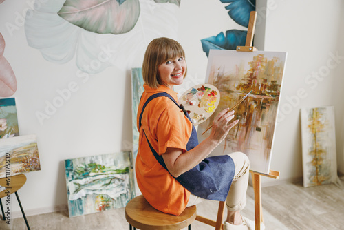 Full body back view elderly artist woman 50 years old wear casual clothes sitting near easel with painting artwork paint look camera spend free spare time in living room indoor. Leisure hobby concept.
