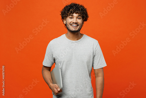 Side view young cheerful fun happy Indian man wears t-shirt casual clothes hold closed laptop pc computer browsing internet isolated on orange red color background studio portrait. Lifestyle concept.