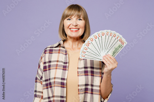 Elderly blonde happy woman 50s years old wear beige t-shirt shirt casual clothes hold in hand fan of cash money in dollar banknotes isolated on plain pastel light purple background. Lifestyle concept.