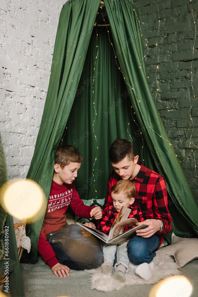 Childs enjoying time together. Merry Christmas. Children reading books for little brother on floor. Large big family with three kids. Interior with canopy with garlands. Concept of winter holiday.