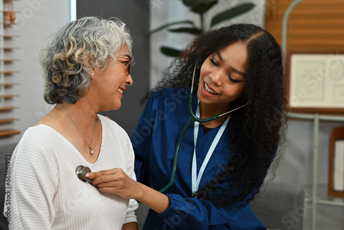 An African-Asian doctor wearing a scrub uniform listening to an older woman lung or heart sounds with a stethoscope during a visit at home, medical checkup and health care concept