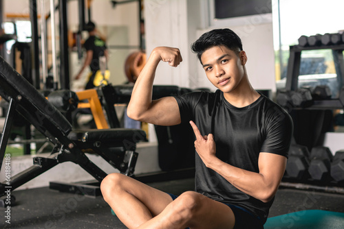 A young handsome and confident asian man flexing his bicep while sitting on the mat at the gym.
