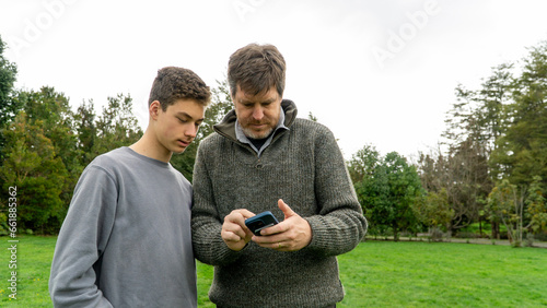 Father and teenage son share a moment in the garden, connecting while looking at something on the dad's phone together.