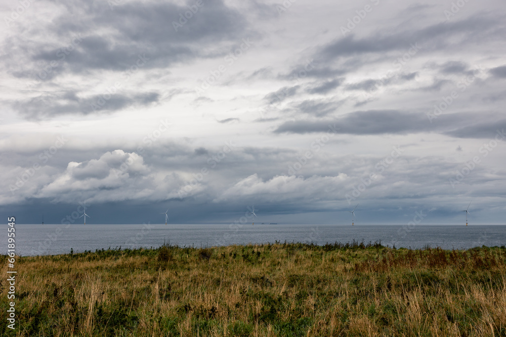 A line of five large offshore windmills, turbines for alternative green, renewable energy, electricity, just of the English coast with cliffs and dramatic clouds.