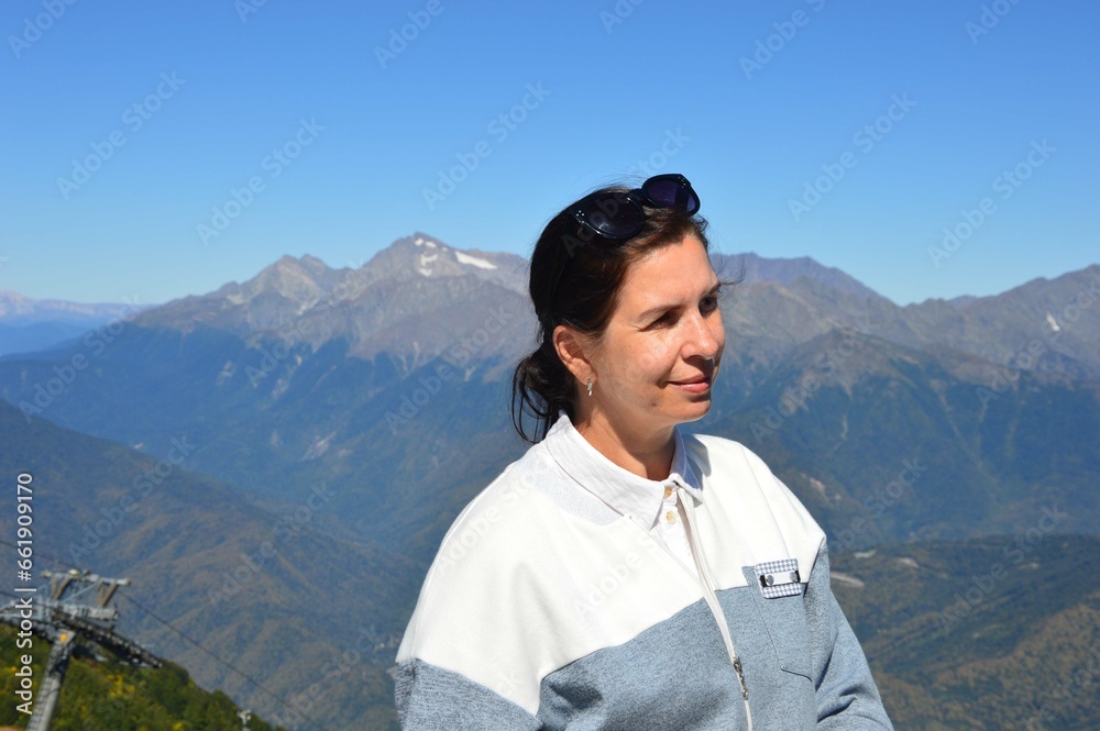 A happy young woman stands against the backdrop of a beautiful mountain landscape. Caucasian brunette enjoys her vacation. Portrait of a female traveler.