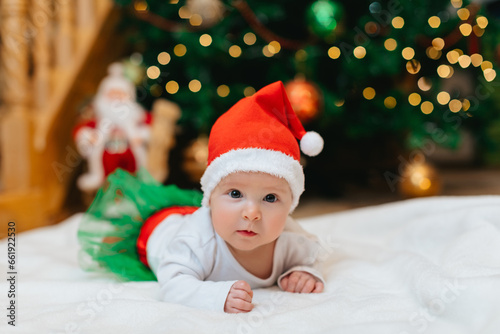 Baby girl with Christmas elf clothes is lying on a white blanket near Christmas tree with bokeh.  Christmas concept. Infant baby in her first Christmas. Funny baby.  © VikaNorm