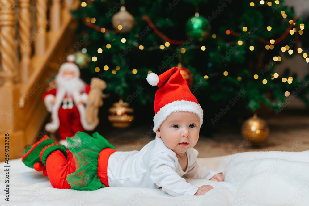 Baby girl with Christmas elf clothes is lying on a white blanket near Christmas tree with bokeh. Christmas concept. Infant baby in her first Christmas. Funny baby. Baby girl with Christmas elf clothes is lying on a white blanket near Christmas tree with bokeh. Christmas concept. Infant baby in her first Christmas. Funny baby.