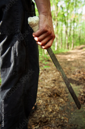Seen from waist-level, a man holds a machete as he walks down a path in the woods.