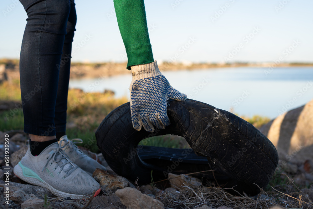 Obraz premium Unrecognizable person wearing gloves picking up a worn vehicle cover from a landfill in a coastal marine estuarine wetland. Photo with space for text.