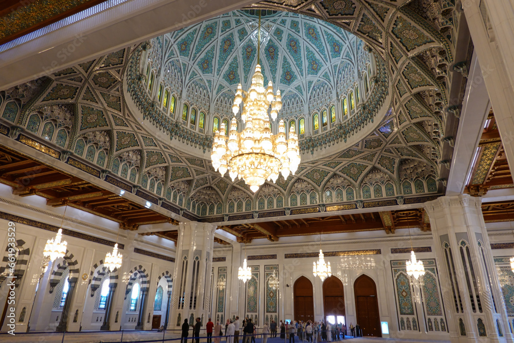 Prayer hall with white marble side colonnade and ornate dome on the ceiling and chandelier in the centre, in the background tourists queuing up at the great mosque of Sultan Qaboos. Oman.