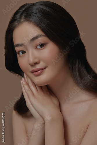 Beautiful young Asian model with perfectly clean and smooth skin folded her hands in the palm of her hand and put it to her face stands on a beige background