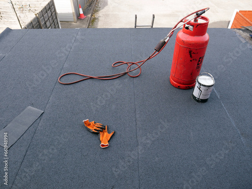 Red propane gas cylinder with connected blowtorch standing on almost finished new bitumen roofing felt covered flat rooftop.