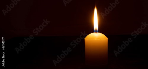 Single burning candle flame or light glowing on a big white candle on black or dark background on table in church for Christmas, funeral or memorial service with copy space.