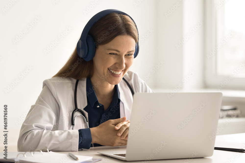 Smiling woman GP in headphones sit at desk give on-line help use laptop, talk to clinic patient, explain medical treatment. Successful therapist take part in briefing via videoconference. E-health app Smiling woman GP in headphones sit at desk give on-line help use laptop, talk to clinic patient, explain medical treatment. Successful therapist take part in briefing via videoconference. E-health app