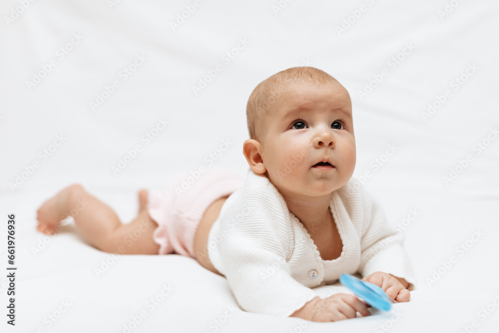 Infant baby girl in a white knitted blouse and pink knitted underpants is lying on stomach on a white blanket and playing with teething toy. Close up. Infant baby girl in a white knitted blouse and pink knitted underpants is lying on stomach on a white blanket and playing with teething toy. Close up.