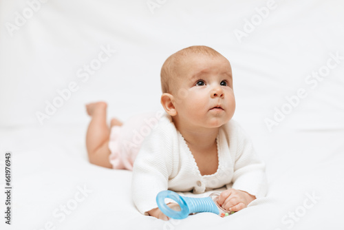 Infant baby girl in a white knitted blouse and pink knitted underpants is lying on stomach on a white blanket and playing with teething toy. Close up. 