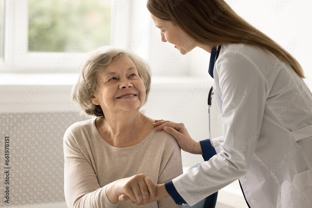 Obraz premium Young female doctor in uniform support old patient hold her palm, give hope, congratulate with good treatment result. Happy aged lady visit therapist for health check up. Professional medical services