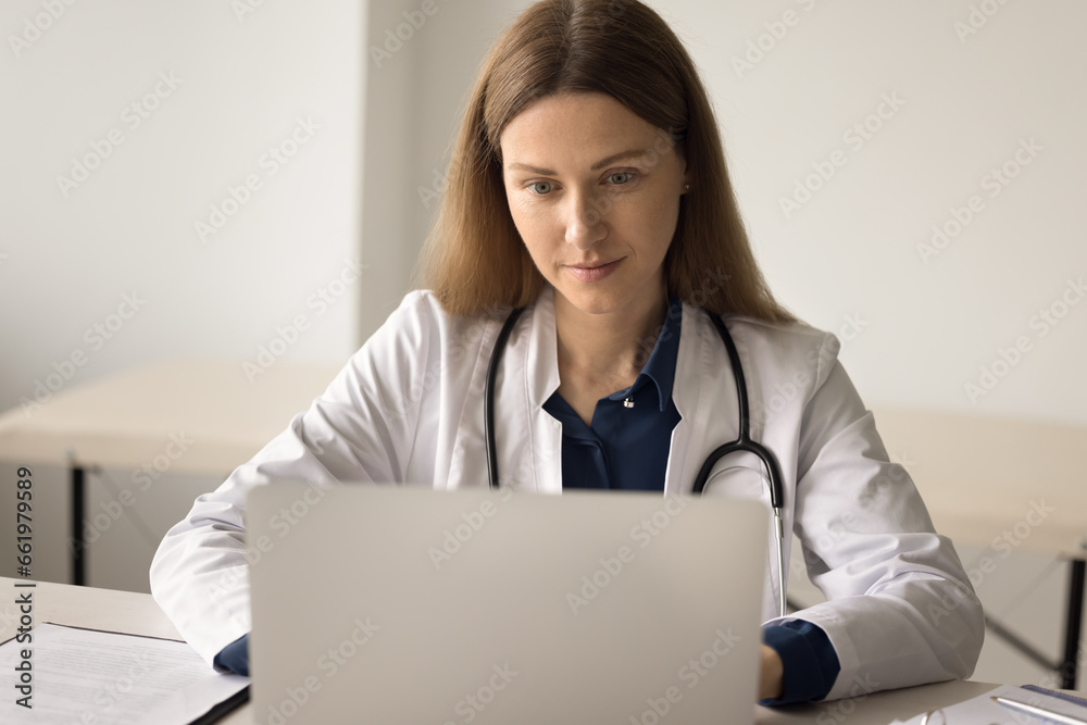 Attractive focused young female doctor in uniform sit at desk work on laptop online, use new medical application. Serious therapist consult patient remotely, give prescription by internet. Healthcare Attractive focused young female doctor in uniform sit at desk work on laptop online, use new medical application. Serious therapist consult patient remotely, give prescription by internet. Healthcare