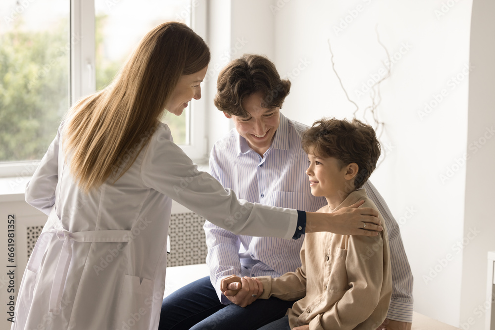 Fototapeta premium Young female pediatrician in white coat support little boy patient touch kid shoulder, give advice, provides medical assistance. Happy small child enjoy checkup meeting with dad. Check-up, healthcare