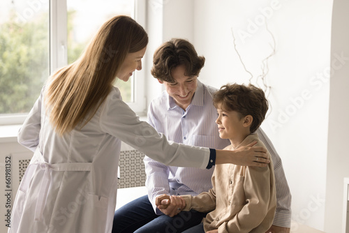 Young female pediatrician in white coat support little boy patient touch kid shoulder, give advice, provides medical assistance. Happy small child enjoy checkup meeting with dad. Check-up, healthcare