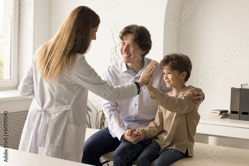 Happy little boy visited doctor in clinic with caring dad. Overjoyed female pediatrist gives high five to cute boy after check up in hospital, celebrate successful treatment results. Medical services