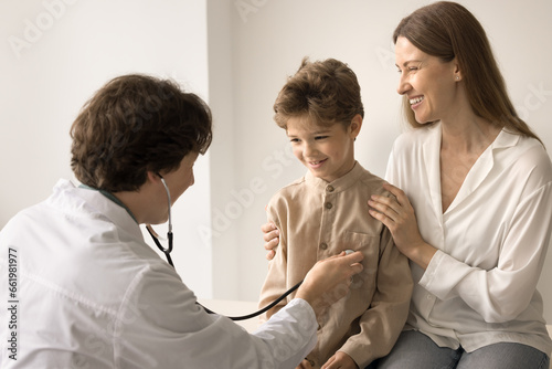 Cute little kid with mother visit doctor for health check. Caring man therapist in white uniform examining child, listen heart with stethoscope, do medical healthcare. Cardiology, childcare, medicine