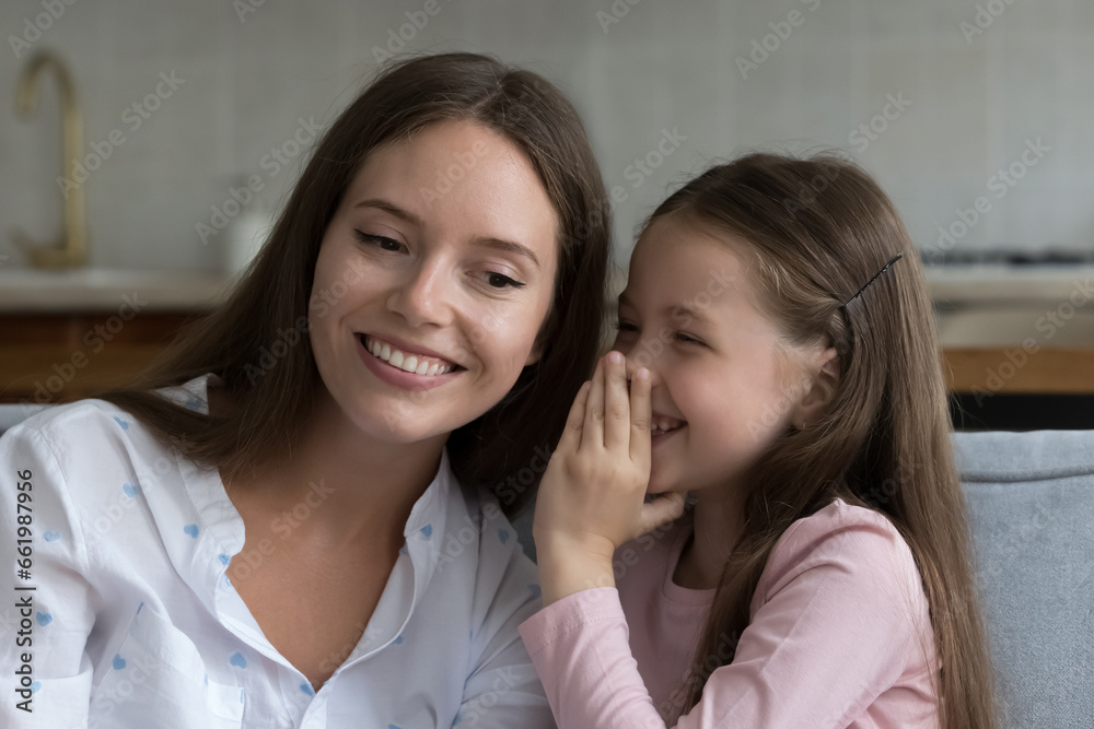 Cheerful excited little girl sharing secret with happy mom, whispering in ear, laughing. Mother and cute daughter kid enjoying being friends, trust, family leisure time, talking, resting on home sofa Cheerful excited little girl sharing secret with happy mom, whispering in ear, laughing. Mother and cute daughter kid enjoying being friends, trust, family leisure time, talking, resting on home sofa