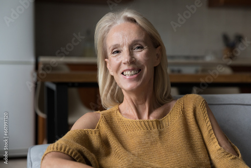 Happy blonde senior 60s woman smiling at camera, looking, speaking at webcam, talking on video call, resting on home couch in living room. Screen view, home head shot portrait. Elderly age concept