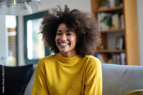 Radiant woman with voluminous curly hair smiles in a cozy living room setting, adorned in a yellow sweater.