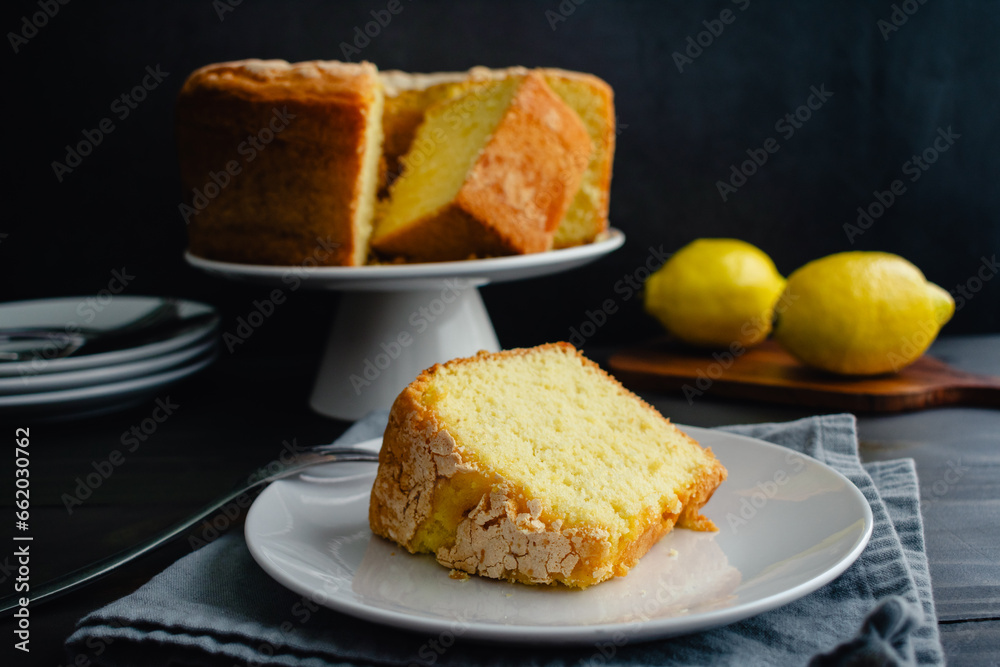 Lemon Pound Cake Slice on a Dessert Plate with a Fork: Plated slice of lemon pound cake with a whole cake on cake stand in the background Lemon Pound Cake Slice on a Dessert Plate with a Fork: Plated slice of lemon pound cake with a whole cake on cake stand in the background