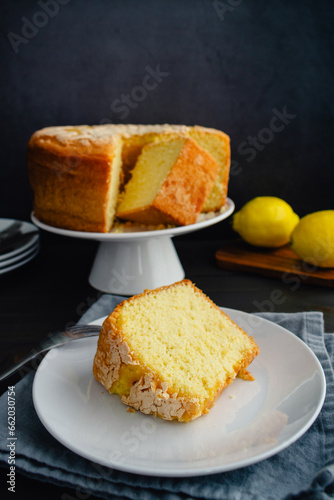 Lemon Pound Cake Slice on a Dessert Plate with a Fork: Plated slice of lemon pound cake with a whole cake on cake stand in the background