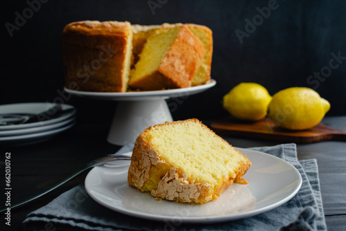 Lemon Pound Cake Slice on a Dessert Plate with a Fork: Plated slice of lemon pound cake with a whole cake on cake stand in the background