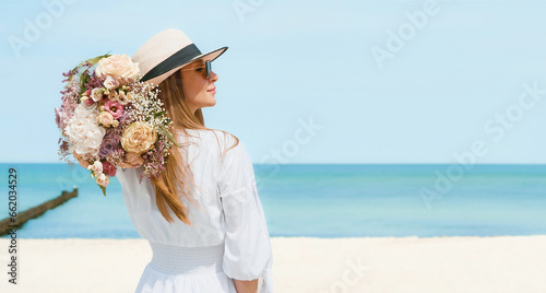 Joyful Young Woman Holding a Bouquet of Flowers on a Beach with Blue Sky Background