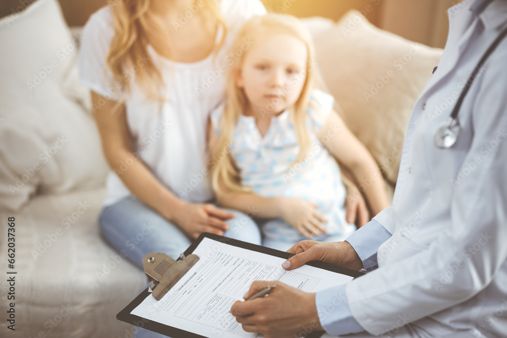 Doctor and patient. Pediatrician using clipboard while examining little girl with her mother at home. Sick and unhappy child at medical exam Doctor and patient. Pediatrician using clipboard while examining little girl with her mother at home. Sick and unhappy child at medical exam