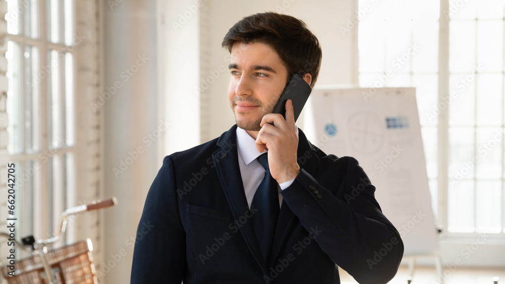 Happy pensive business owner man in formal suite and tie talking on cell in office, looking away, standing in office room with whiteboard behind, making call, smiling, enjoying conversation Happy pensive business owner man in formal suite and tie talking on cell in office, looking away, standing in office room with whiteboard behind, making call, smiling, enjoying conversation