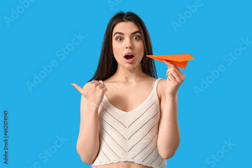 Surprised young woman with paper plane pointing at something on blue background
