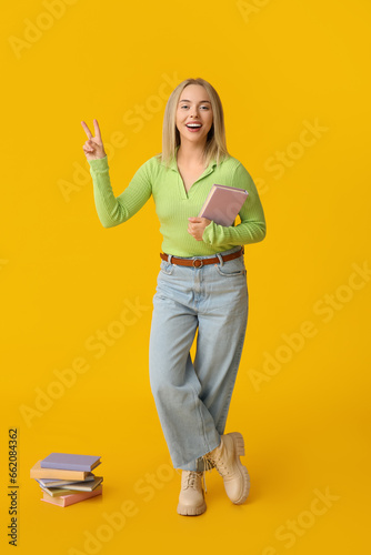 Beautiful young happy woman with many books showing victory gesture on yellow background