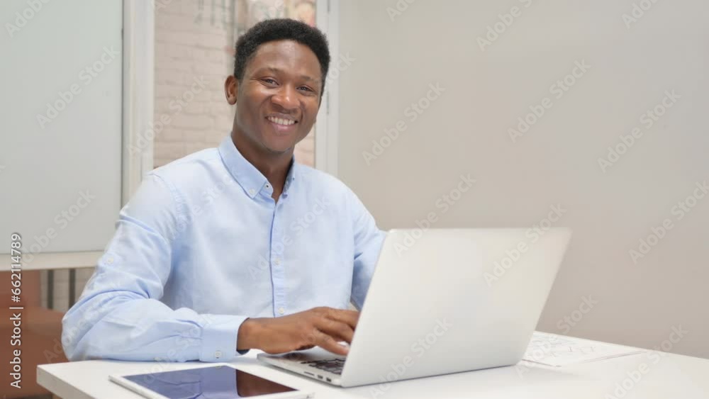 African Businessman Smiling at Camera while Working on Laptop in Office African Businessman Smiling at Camera while Working on Laptop in Office