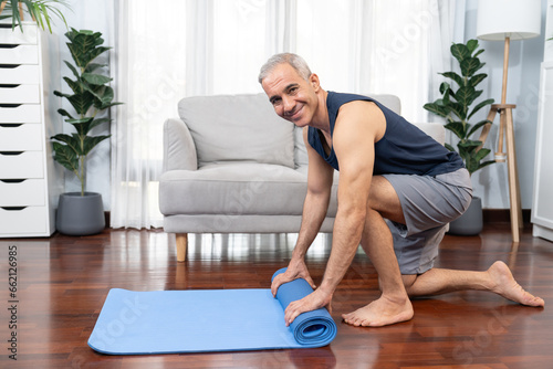 Active and sporty senior man preparing, rolling fitness exercising mat on living room floor at home. Home exercise as concept of healthy fit body lifestyle after retirement. Clout