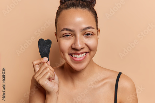 Positive Latin woman making face massage with jade gua sha in morning face care routine smiles happily undergoes anti aging procedure at home poses indoor. Rejuvenation beauty and wellness concept
