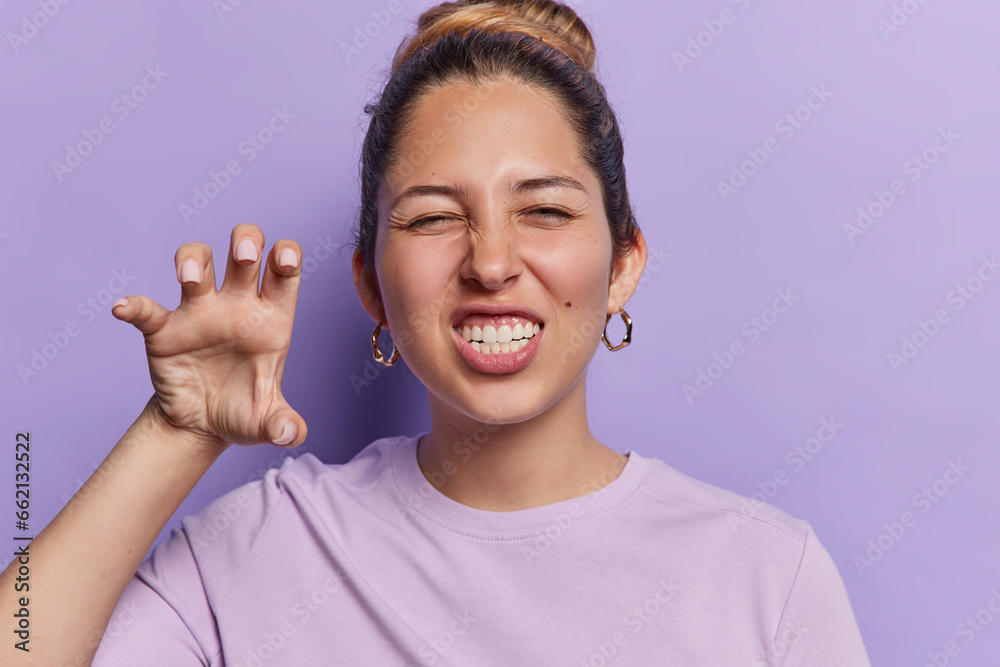 Photo of scary European woman making cat claws gesture with hands raised clenches teeth pretends being animal and roars dressed casually poses against purple background. Body language concept Photo of scary European woman making cat claws gesture with hands raised clenches teeth pretends being animal and roars dressed casually poses against purple background. Body language concept