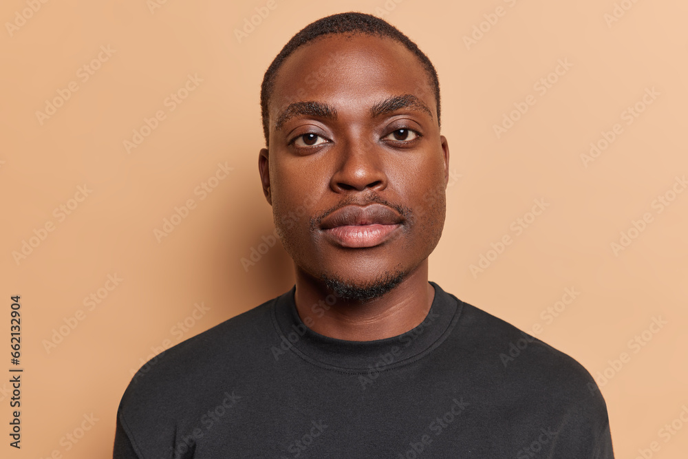 Obraz premium People and positive emotions concept. Indoor close up of young happy satisfied African american male standing in centre isolated on beige background looking straight at camera dressed in black t shirt