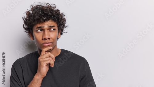 Studio portrait of young thoughtful Hindu man standing on left on white background in black t shirt keeping hand on chin with blank space for your advertisement trying to solve difficult problem