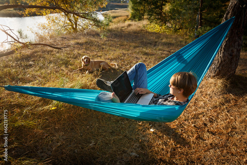 Young woman in hammok using laptop working outdoor facing lake on a sunset together with her home pet dog.