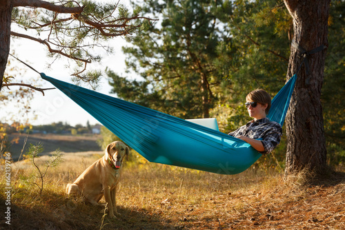 Young woman in hammok using laptop working outdoor facing lake on a sunset together with her home pet dog.