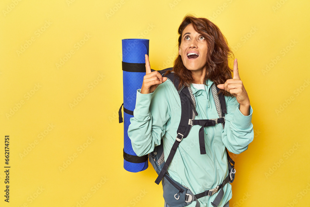 Woman with hiking backpack and mat on yellow pointing upside with opened mouth.
