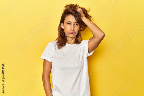 Middle-aged woman on a yellow backdrop tired and very sleepy keeping hand on head.
