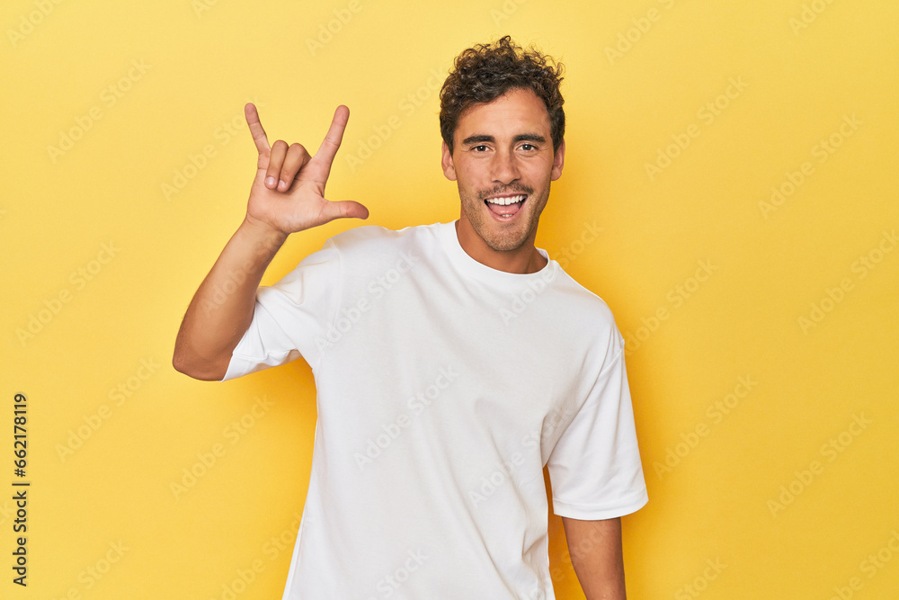 Young Latino man posing on yellow background showing a horns gesture as a revolution concept. Young Latino man posing on yellow background showing a horns gesture as a revolution concept.