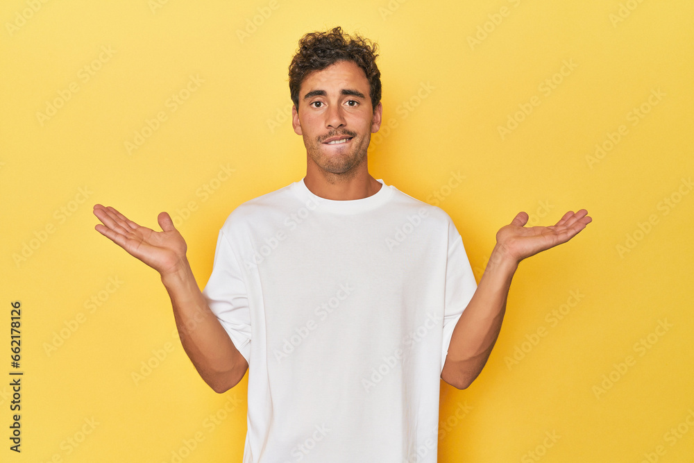Young Latino man posing on yellow background confused and doubtful shrugging shoulders to hold a copy space. Young Latino man posing on yellow background confused and doubtful shrugging shoulders to hold a copy space.