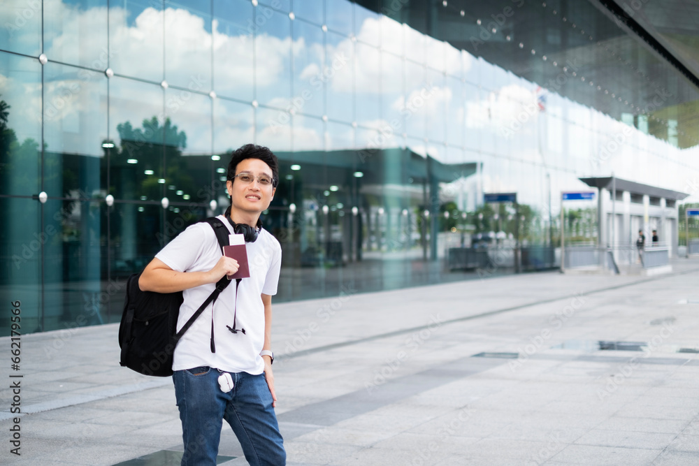 Obraz premium Young Asian millenael man standing with smile in front of air port ready for trip travel alone with passport, black backpack and headphone.